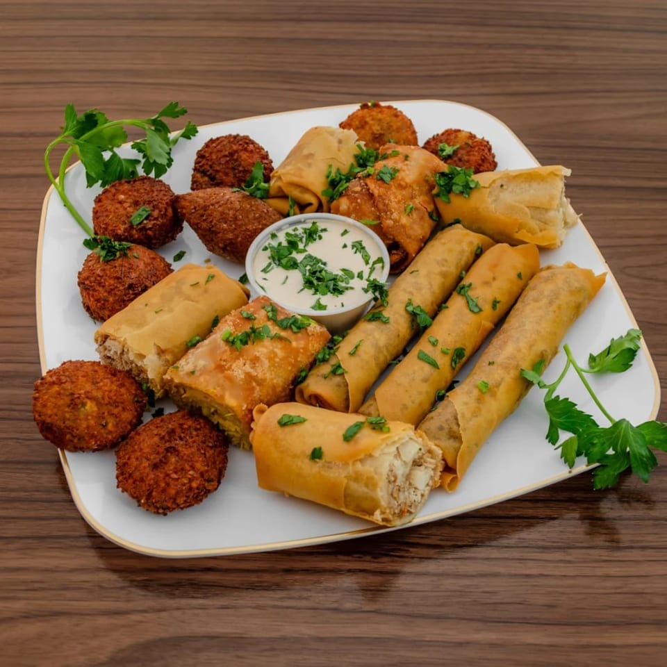 A white platter filled with golden-brown falafel, spring rolls, and a small cup of white dipping sauce.