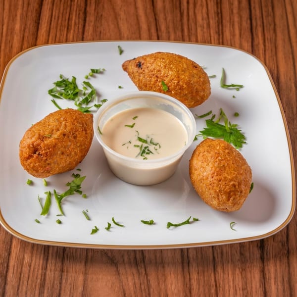 Three golden-brown fried kibbeh on a white plate with a side of tahini sauce and parsley.